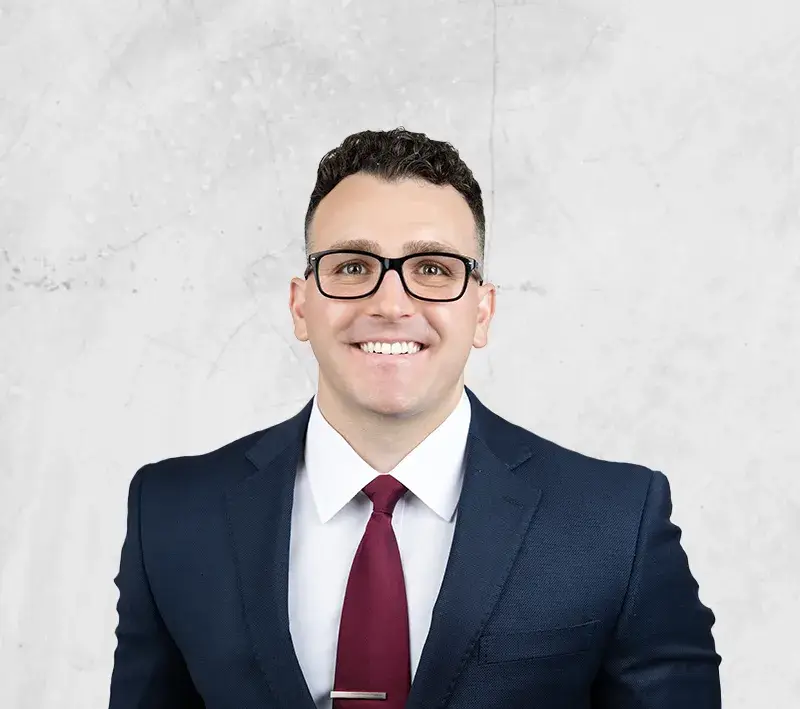 Smiling man in a navy suit, white shirt, burgundy tie and glasses, posing for a professional headshot against a light textured background.