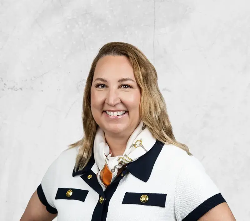 Smiling woman in a white cardigan with navy trim and scarf, posing against a light textured background.