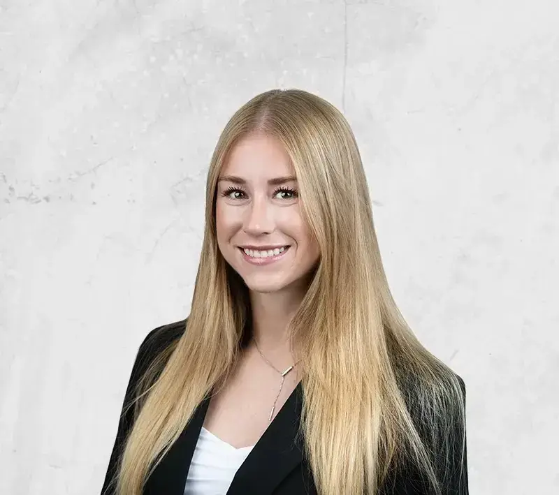 Smiling young woman with long blond hair in a black blazer against a light gray background (professional headshot)