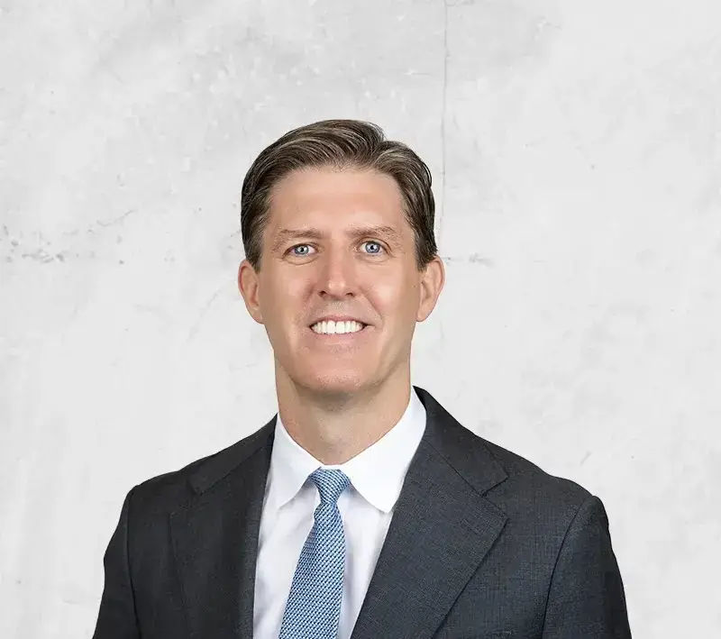 Professional headshot: smiling man in a dark suit and light blue tie against a light gray textured background.
