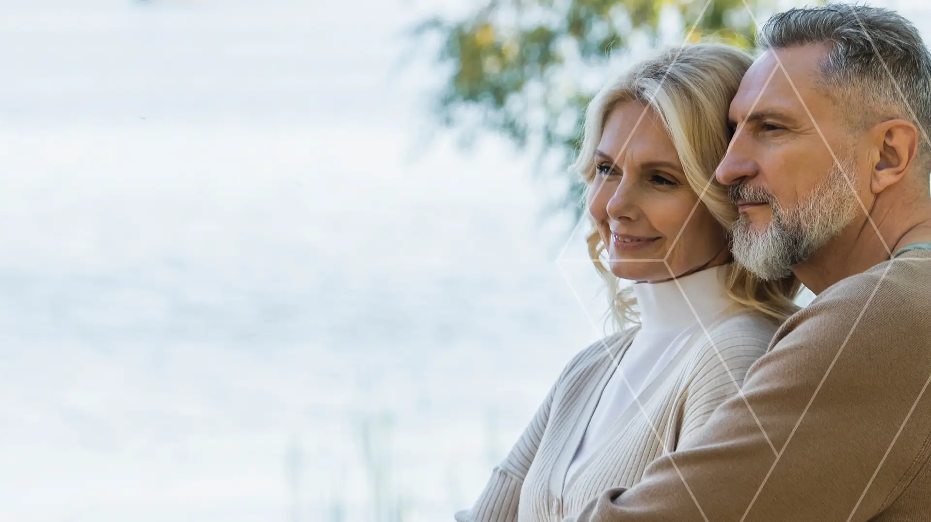 A smiling middle-aged couple stands close together by a lake, with trees in the background.
