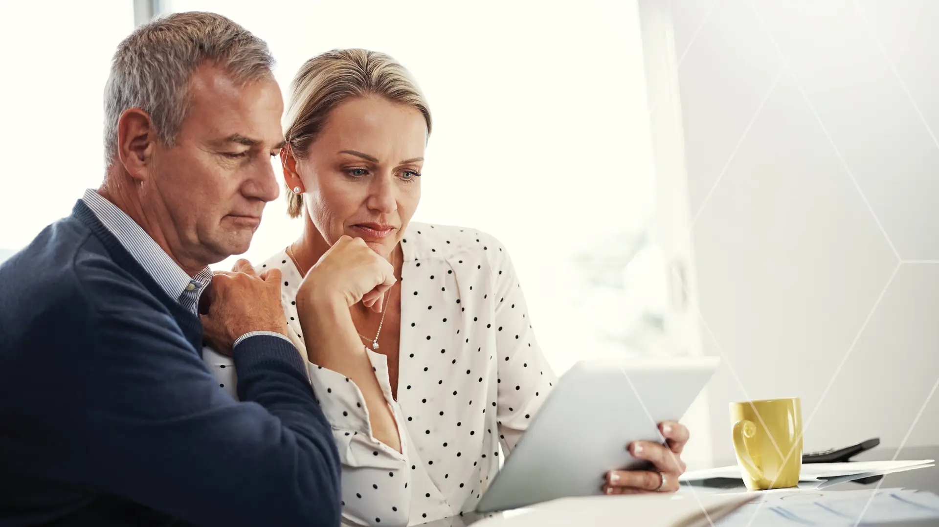Two professionals, a man and a woman, closely review content on a tablet at a bright office desk with a yellow mug nearby.