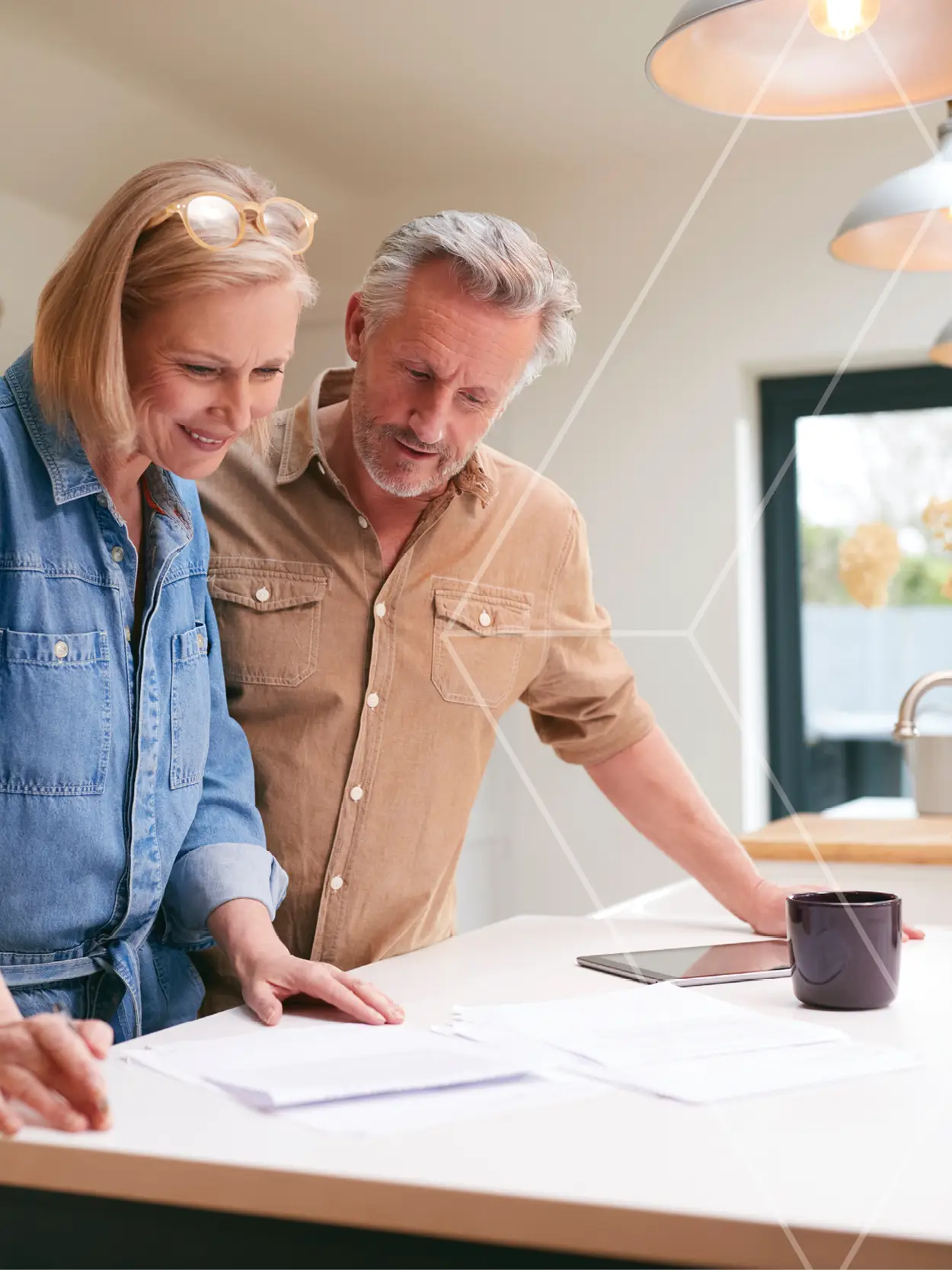 Couple of older adults reviewing documents at a bright kitchen counter with a tablet and mug nearby.