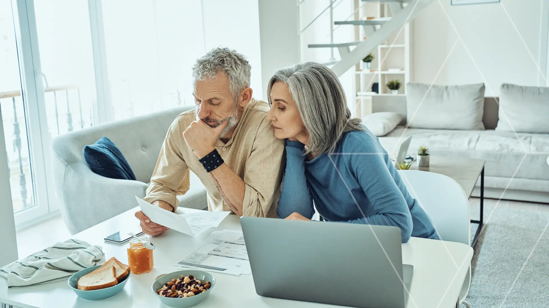Older couple sits at a white table in a bright living room, reading documents with a laptop nearby.