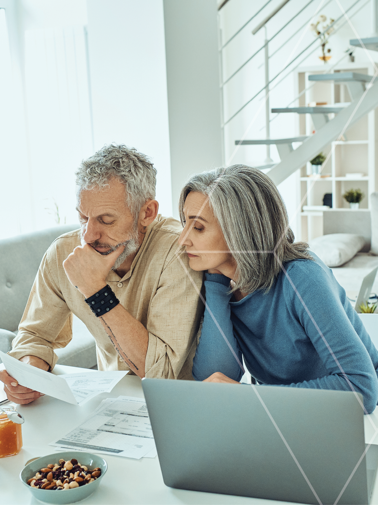 Older couple sitting at a bright white table, reviewing documents with a laptop nearby, looking thoughtful and focused.